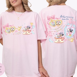 May include: A pale pink t-shirt with a playful design of cartoon characters, teacups, and bows. The front features a heart-shaped frame with characters, a genie lamp, and a teacup. The back of the shirt has the text "Girls just wanna have fun."