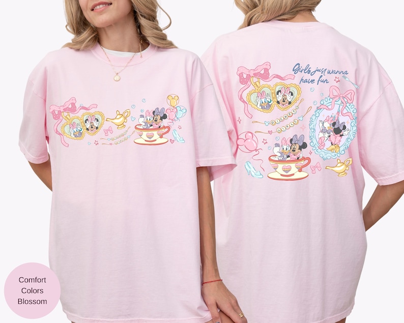 May include: A pale pink t-shirt with a playful design of cartoon characters, teacups, and bows. The front features a heart-shaped frame with characters, a genie lamp, and a teacup. The back of the shirt has the text "Girls just wanna have fun."