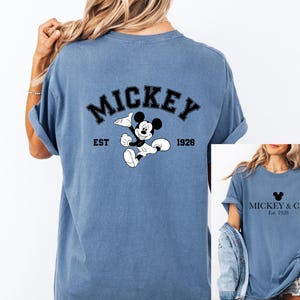 Puede incluir: Dos camisetas azules con un dise&ntilde;o de Mickey Mouse. La parte posterior de una camiseta dice "Mickey Est. 1928" con un dibujo animado de Mickey Mouse. La parte delantera de la otra camiseta dice "Mickey & Co. Est. 1928" con una cabeza de Mickey Mouse.