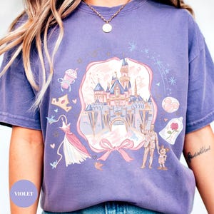 May include: A violet-colored t-shirt featuring a whimsical illustration of a fairytale castle, a dress, a crown, and other themed elements. The design includes a bow and a couple with a child. The word "VIOLET" is printed on the bottom left.