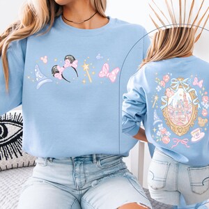May include: Light blue sweatshirt featuring whimsical illustrations. The front displays a wizard hat, Minnie Mouse ears, a magic wand, and a bow. The back showcases a castle within a decorative frame, surrounded by floral and bow accents. A charming and playful design.