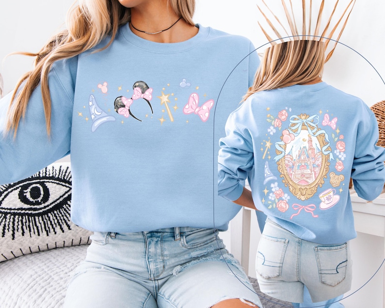 May include: Light blue sweatshirt featuring whimsical illustrations. The front displays a wizard hat, Minnie Mouse ears, a magic wand, and a bow. The back showcases a castle within a decorative frame, surrounded by floral and bow accents. A charming and playful design.