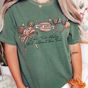 Może przedstawiać: Zielona koszulka z grafiką Złomka z filmu Auta, r&oacute;żową kokardą i napisem "Like Tuh-Mater, But without the Tuh." Projekt zawiera uszy Myszki Miki i logo "Tow Mater Towing & Salvage".