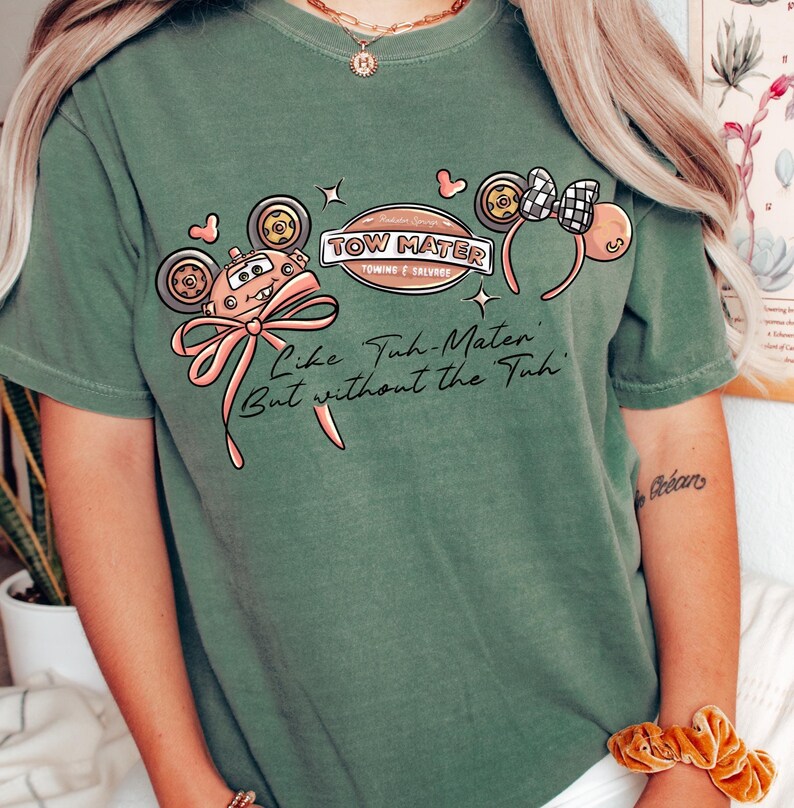 Może przedstawiać: Zielona koszulka z grafiką Złomka z filmu Auta, r&oacute;żową kokardą i napisem "Like Tuh-Mater, But without the Tuh." Projekt zawiera uszy Myszki Miki i logo "Tow Mater Towing & Salvage".