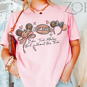 Może przedstawiać: Jasnor&oacute;żowa koszulka z kresk&oacute;wkowym wzorem. Wz&oacute;r zawiera tekst "TOW MATER" i "Like Tuh-Mater But without the Tuh", samoch&oacute;d z kresk&oacute;wek, uszy Myszki Miki i kokardę.