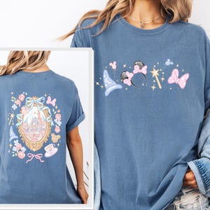 May include: A blue t-shirt featuring a whimsical design with a castle, a magic wand, and mouse ears. The back of the shirt has a framed castle design with floral and bow accents. The shirt is made of a soft material.