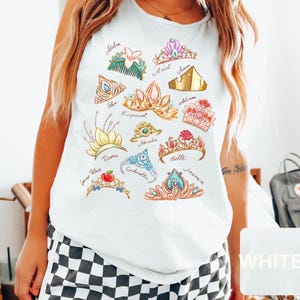 May include: White tank top featuring watercolor illustrations of various princess crowns, each labeled with a name. The crowns are in shades of gold, pink, blue, and green. The shirt is worn by a person with blonde hair and a tattoo on their arm.