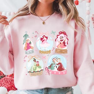 May include: A light pink sweatshirt featuring a design of several snow globes. Each globe contains a princess illustration. The design includes candy canes, bows, and decorative elements, creating a festive and whimsical aesthetic.