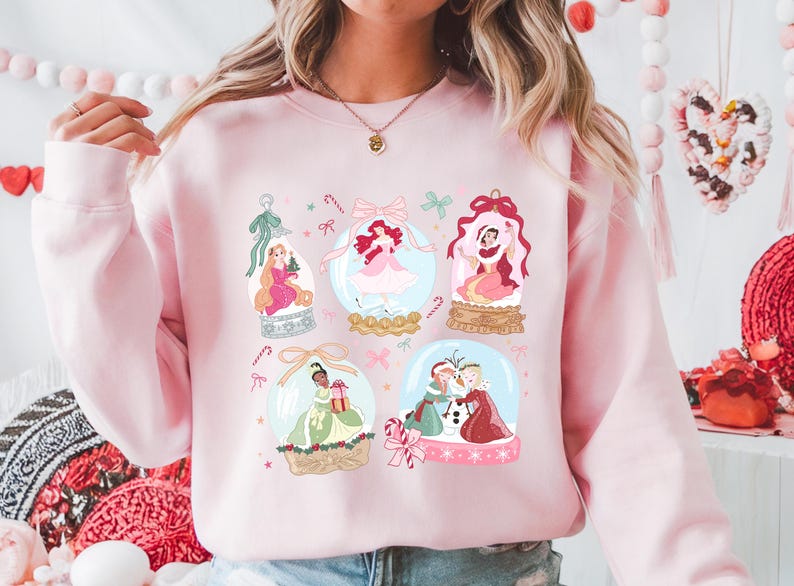 May include: A light pink sweatshirt featuring a design of several snow globes. Each globe contains a princess illustration. The design includes candy canes, bows, and decorative elements, creating a festive and whimsical aesthetic.