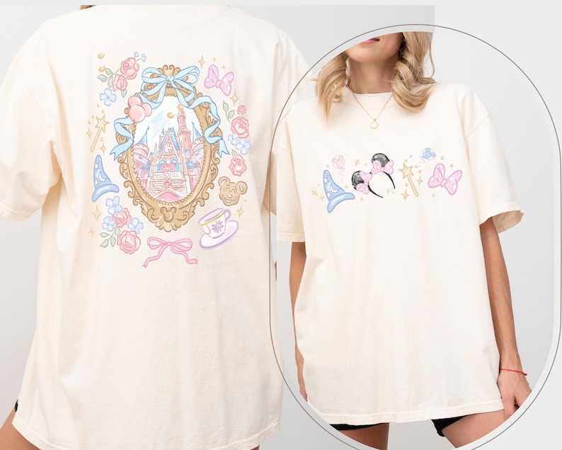 May include: Cream-colored t-shirt featuring a whimsical design. The front showcases a Minnie Mouse ear headband, a magic wand, and a wizard hat. The back displays a castle scene within a decorative frame, surrounded by roses, bows, and a teacup. The design is in pastel colors.