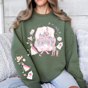 May include: Olive green sweatshirt featuring a castle graphic with pink and blue accents. The design includes a pink bow, dresses, and a rose in a glass dome. The sleeve has additional graphics of dresses and a rose.