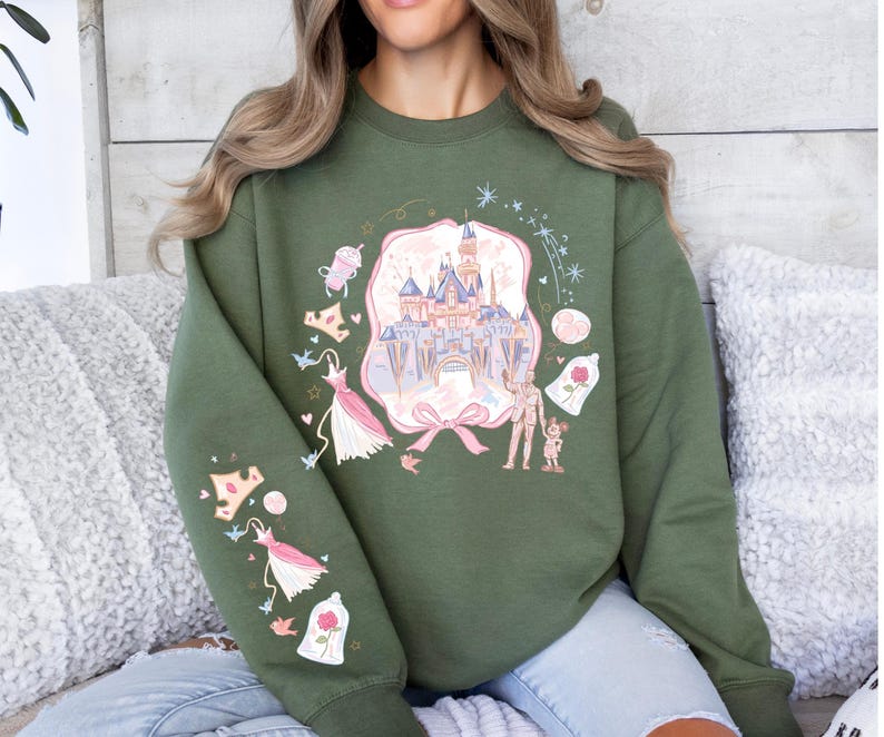 May include: Olive green sweatshirt featuring a castle graphic with pink and blue accents. The design includes a pink bow, dresses, and a rose in a glass dome. The sleeve has additional graphics of dresses and a rose.
