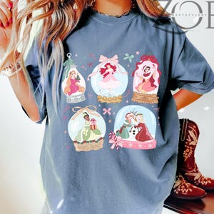 May include: A blue t-shirt featuring six snow globes with princess illustrations. Each globe has a different princess, with pink, red, and green accents. The shirt has the text "ZOE" in the upper right corner.