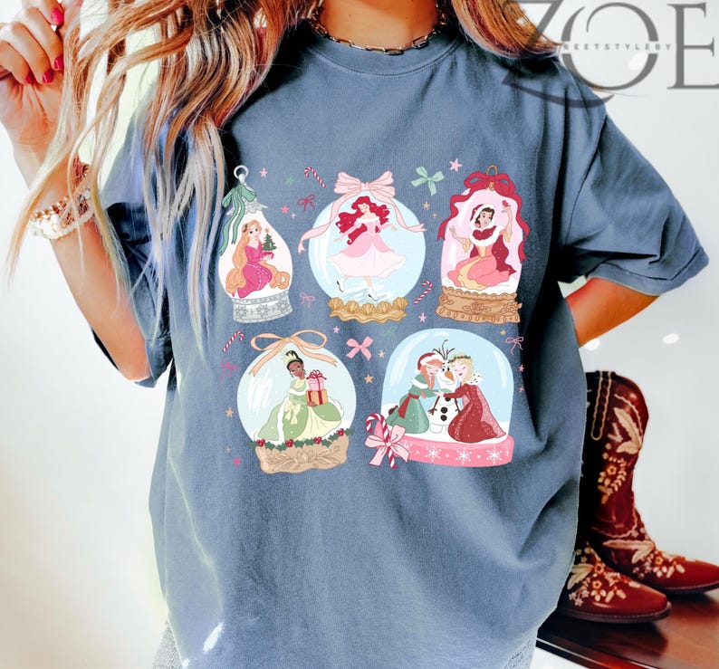 May include: A blue t-shirt featuring six snow globes with princess illustrations. Each globe has a different princess, with pink, red, and green accents. The shirt has the text "ZOE" in the upper right corner.