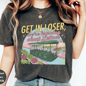 Puede incluir: Una camiseta gris carb&oacute;n con un estampado gr&aacute;fico de un paseo en barco por un invernadero con el texto "GET IN LOSER, we're learning about Lettuce" en letras amarillas y rosas, rodeado de ilustraciones de varias plantas de lechuga.