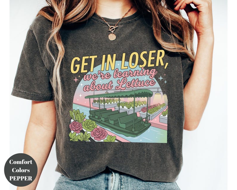 Puede incluir: Una camiseta gris carb&oacute;n con un estampado gr&aacute;fico de un paseo en barco por un invernadero con el texto "GET IN LOSER, we're learning about Lettuce" en letras amarillas y rosas, rodeado de ilustraciones de varias plantas de lechuga.