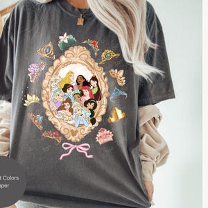 May include: Dark grey t-shirt featuring a gold-framed portrait of Disney princesses. Surrounding the portrait are various tiaras and floral accents. The text "Comfort Colors Pepper" is displayed on a circular label.
