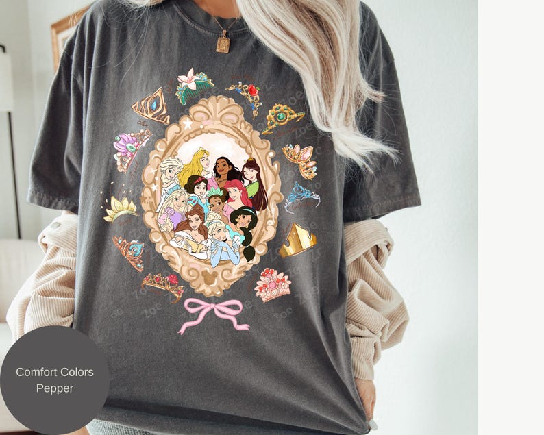 May include: Dark grey t-shirt featuring a gold-framed portrait of Disney princesses. Surrounding the portrait are various tiaras and floral accents. The text "Comfort Colors Pepper" is displayed on a circular label.