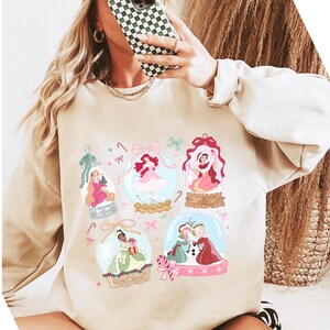 May include: Beige sweatshirt featuring colorful illustrations of princesses inside snow globes. The design includes pink, green, and blue accents, with candy cane and bow details. The sweatshirt has long sleeves and a crew neck.