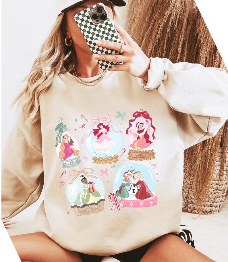 May include: Beige sweatshirt featuring colorful illustrations of princesses inside snow globes. The design includes pink, green, and blue accents, with candy cane and bow details. The sweatshirt has long sleeves and a crew neck.