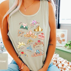May include: Sandstone-colored tank top featuring colorful illustrations of various princess tiaras. Each tiara is uniquely designed with different colors and embellishments. The names of the princesses are written below each tiara. The tank top is made of a soft material.