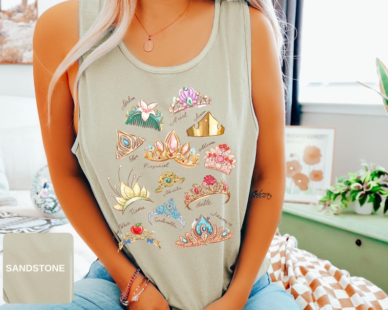 May include: Sandstone-colored tank top featuring colorful illustrations of various princess tiaras. Each tiara is uniquely designed with different colors and embellishments. The names of the princesses are written below each tiara. The tank top is made of a soft material.