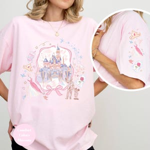 May include: A light pink t-shirt with a whimsical castle graphic, featuring a castle, characters, and other elements. The sleeve has additional embroidered details. The shirt has the words "Comfort Colors Blossom" on the bottom.