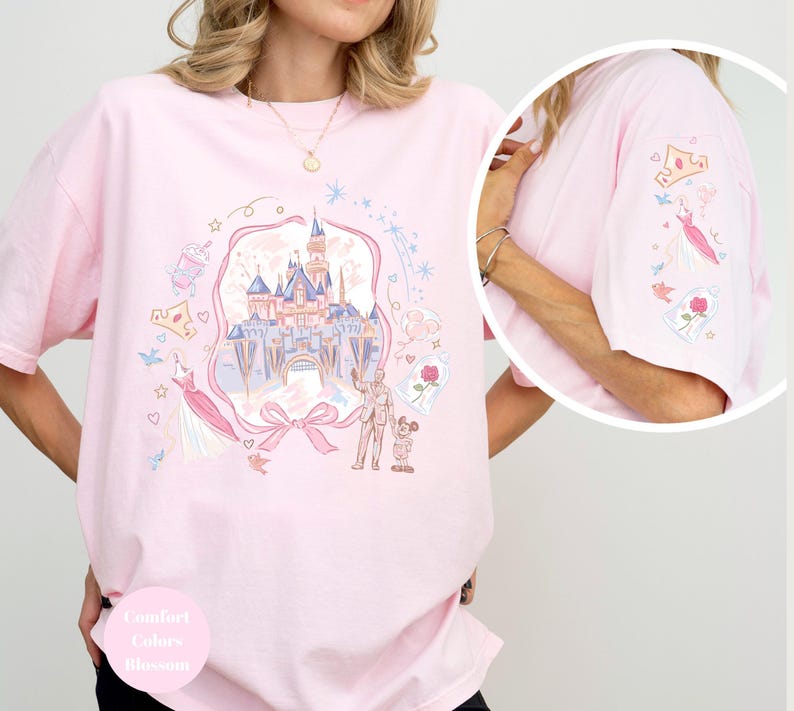 May include: A light pink t-shirt with a whimsical castle graphic, featuring a castle, characters, and other elements. The sleeve has additional embroidered details. The shirt has the words "Comfort Colors Blossom" on the bottom.
