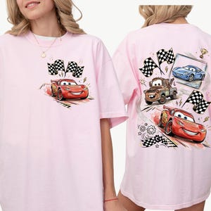 Puede incluir: Camiseta rosa claro con un dise&ntilde;o de dibujos animados de un coche de carreras rojo y banderas a cuadros. La parte trasera de la camiseta muestra m&aacute;s coches de carreras y banderas. El estilo es de dibujos animados.