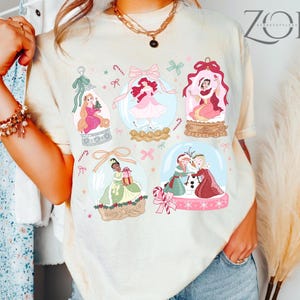 May include: Cream-colored t-shirt featuring illustrations of princesses inside snow globes. The design includes pink bows, candy canes, and stars. The shirt has a relaxed fit and a crew neck.