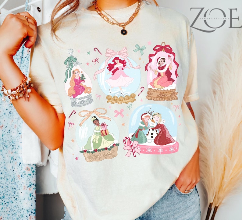 May include: Cream-colored t-shirt featuring illustrations of princesses inside snow globes. The design includes pink bows, candy canes, and stars. The shirt has a relaxed fit and a crew neck.