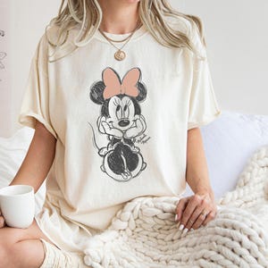 May include: A white t-shirt with a black and white illustration of Minnie Mouse. Minnie Mouse is wearing a pink bow and has her hands on her cheeks. The t-shirt has a distressed look.