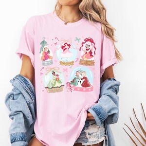 May include: Pink t-shirt featuring six princess-themed snow globes with candy cane and bow accents. The snow globes contain illustrations of princesses in various outfits. The shirt is worn by a person.