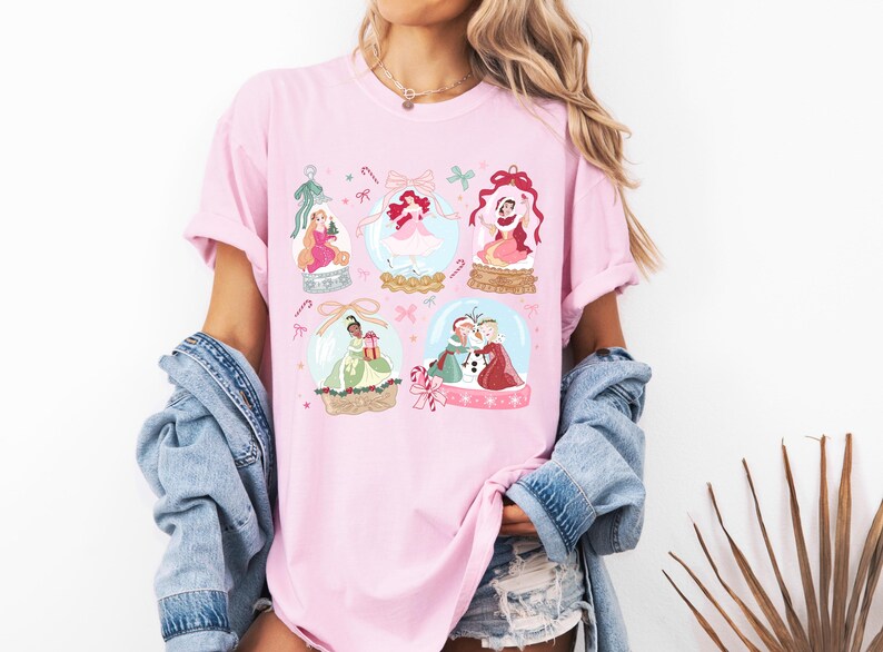 May include: Pink t-shirt featuring six princess-themed snow globes with candy cane and bow accents. The snow globes contain illustrations of princesses in various outfits. The shirt is worn by a person.