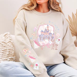 May include: Beige sweatshirt with a colorful illustration of a fairytale castle, dresses, and other themed graphics. The design is framed by a pink ribbon. The sleeves feature additional matching graphics. The sweatshirt is a casual, comfortable style.