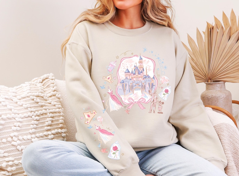 May include: Beige sweatshirt with a colorful illustration of a fairytale castle, dresses, and other themed graphics. The design is framed by a pink ribbon. The sleeves feature additional matching graphics. The sweatshirt is a casual, comfortable style.