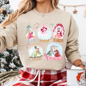 May include: A beige sweatshirt featuring six snow globes with princess illustrations. Each globe has a different princess, with pink and green accents. The sweatshirt is paired with red and white plaid pajama pants.
