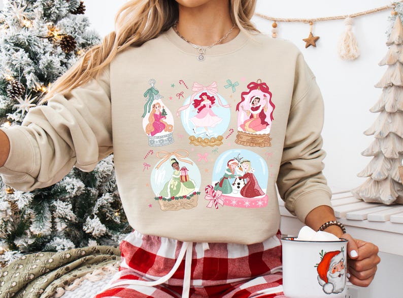 May include: A beige sweatshirt featuring six snow globes with princess illustrations. Each globe has a different princess, with pink and green accents. The sweatshirt is paired with red and white plaid pajama pants.