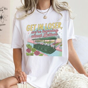 Puede incluir: Una camiseta blanca de manga corta con un estampado gr&aacute;fico que incluye el texto GET IN LOSER, we're learning about Lettuce. La ilustraci&oacute;n muestra un barco verde en un canal rodeado de hileras de plantas y flores rosas bajo un cielo pastel.