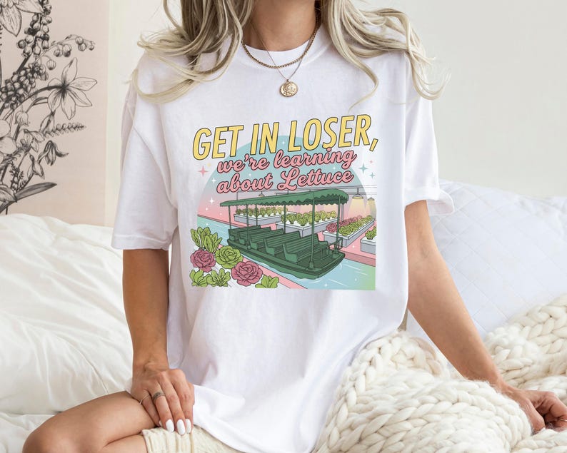 Puede incluir: Una camiseta blanca de manga corta con un estampado gr&aacute;fico que incluye el texto GET IN LOSER, we're learning about Lettuce. La ilustraci&oacute;n muestra un barco verde en un canal rodeado de hileras de plantas y flores rosas bajo un cielo pastel.
