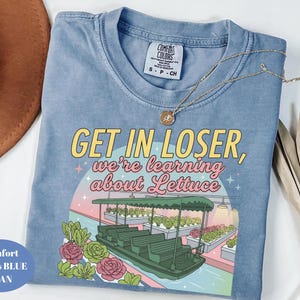 Puede incluir: Una camiseta color azul jean con un estampado gr&aacute;fico que muestra un barco verde sobre el agua, hileras de lechuga y el texto "GET IN LOSER, we're learning about Lettuce" en letras amarillas y rosas. La camiseta tiene cuello redondo y etiqueta talla S.