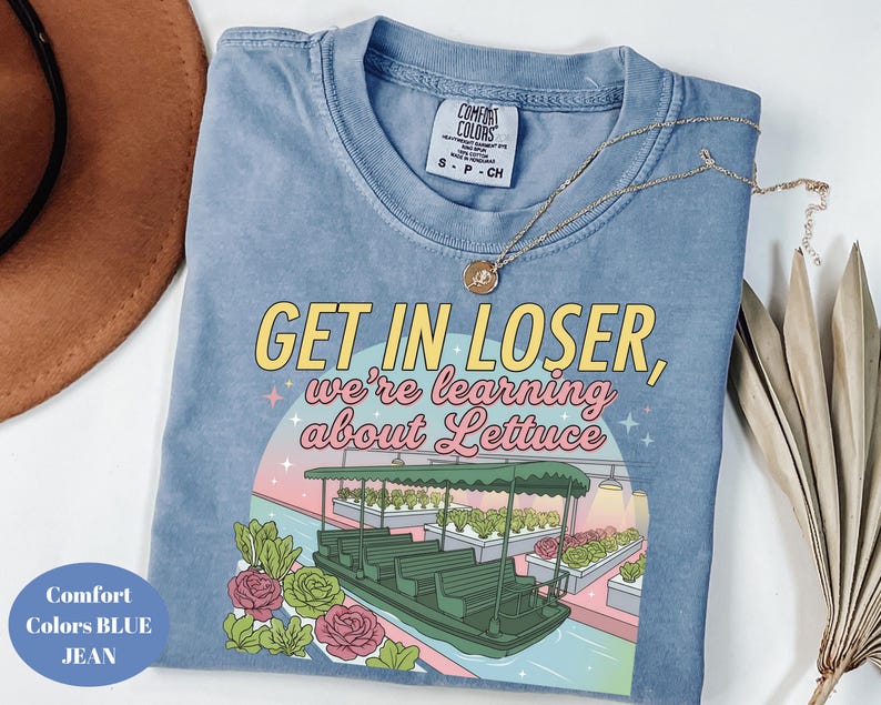 Puede incluir: Una camiseta color azul jean con un estampado gr&aacute;fico que muestra un barco verde sobre el agua, hileras de lechuga y el texto "GET IN LOSER, we're learning about Lettuce" en letras amarillas y rosas. La camiseta tiene cuello redondo y etiqueta talla S.