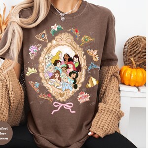May include: Espresso-colored t-shirt featuring a framed illustration of Disney princesses. The frame is surrounded by tiaras and bows. The shirt is part of the Comfort Colors collection.