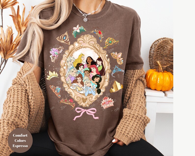 May include: Espresso-colored t-shirt featuring a framed illustration of Disney princesses. The frame is surrounded by tiaras and bows. The shirt is part of the Comfort Colors collection.
