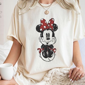 May include: A white t-shirt with a black and red cartoon image of Minnie Mouse. Minnie Mouse is wearing a red bow with white polka dots and has a black and white polka dot dress.