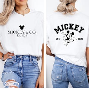 Puede incluir: Camiseta blanca con una cabeza de Mickey Mouse negra y el texto "Mickey & Co. Est. 1928" en la parte delantera. La parte posterior de la camiseta tiene un gr&aacute;fico negro de Mickey Mouse con el texto "Mickey Est. 1928" en un arco negro.