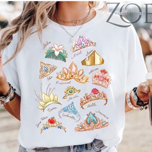 K&ouml;nnte beinhalten: Wei&szlig;es T-Shirt mit farbenfrohen Illustrationen verschiedener Prinzessinnenkronen, jeweils mit Namen wie "Ariel" und "Belle" versehen. Die Kronen, mit Edelsteinen und Blumen, bieten ein verspieltes und k&uuml;nstlerisches Design.