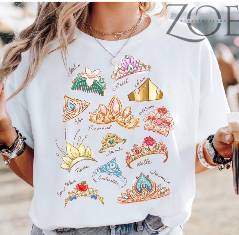 K&ouml;nnte beinhalten: Wei&szlig;es T-Shirt mit farbenfrohen Illustrationen verschiedener Prinzessinnenkronen, jeweils mit Namen wie "Ariel" und "Belle" versehen. Die Kronen, mit Edelsteinen und Blumen, bieten ein verspieltes und k&uuml;nstlerisches Design.