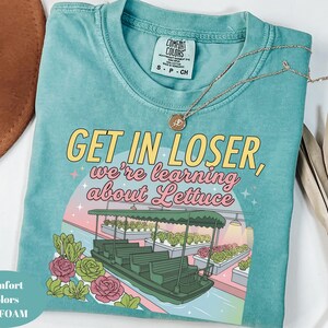 Puede incluir: Una camiseta verde espuma de mar con un gr&aacute;fico de un barco navegando por un invernadero con filas de lechugas. El texto GET IN LOSER, we're learning about Lettuce aparece en letras amarillas y rosas. La camiseta tiene una etiqueta Comfort Colors.