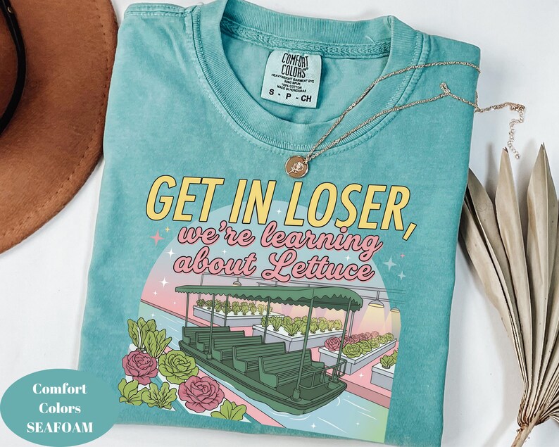 Puede incluir: Una camiseta verde espuma de mar con un gr&aacute;fico de un barco navegando por un invernadero con filas de lechugas. El texto GET IN LOSER, we're learning about Lettuce aparece en letras amarillas y rosas. La camiseta tiene una etiqueta Comfort Colors.